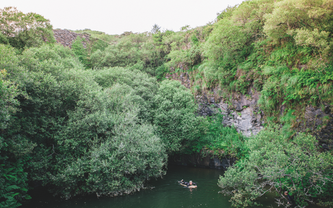 Two people float in a dark green pool surrounded by lush trees and rocky cliffs in a secluded gorge.
