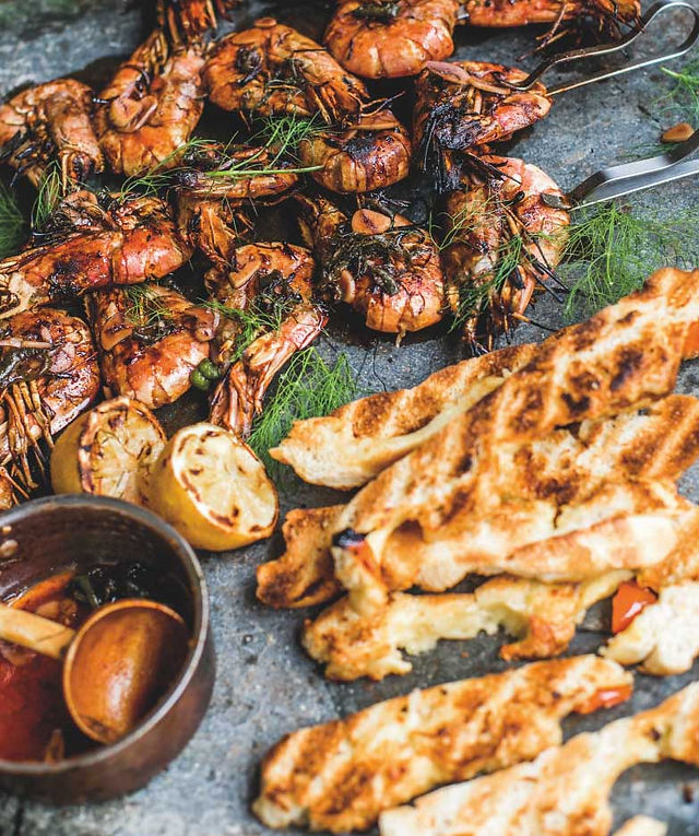 Grilled shrimp with herbs, charred bread, and lemon halves on a rustic stone serving board, with a cozy, appetizing feel