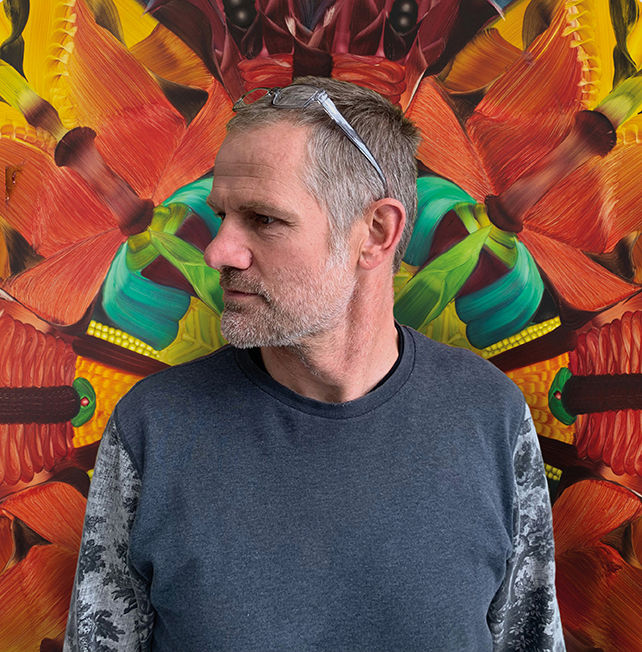 Gray-haired man with glasses on his head looks left in front of a vivid orange, green, and yellow abstract mural. Andy Harper in front of ‘The Carriers’, 2023, oil on board, 120cm (diameter)