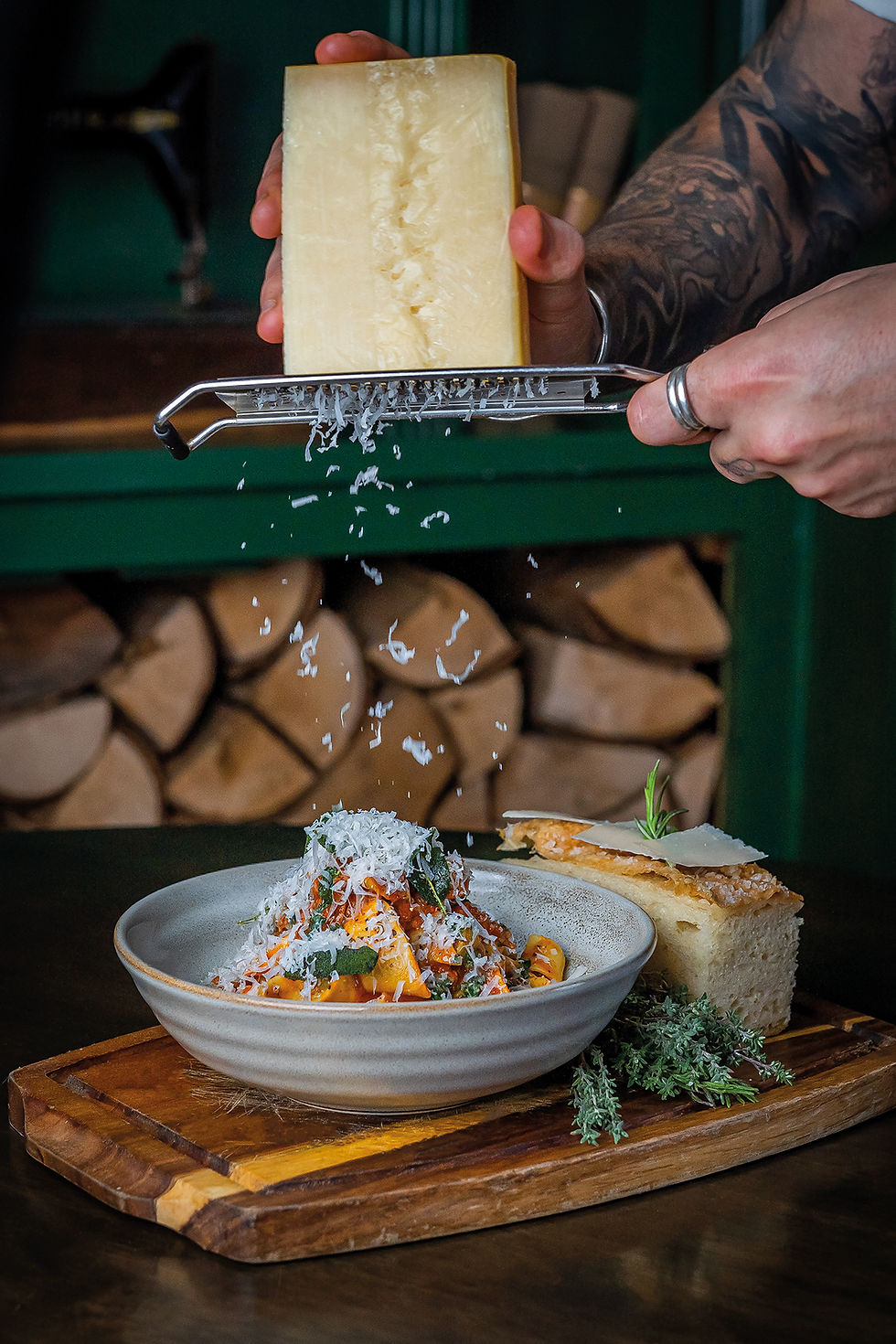 Hands grate cheese over a pasta dish on a wooden board, with bread and herbs in a rustic kitchen. Lamb Ragu recipe