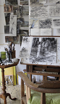 Art studio with charcoal landscape sketches pinned on the wall, an easel holding a forest drawing, and brushes on a side table.