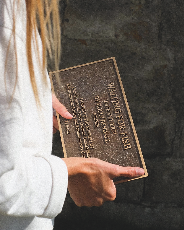 Person in a white sleeve holds a bronze plaque reading WAITING FOR FISH by Holly Bhendall, against a dark stone wall