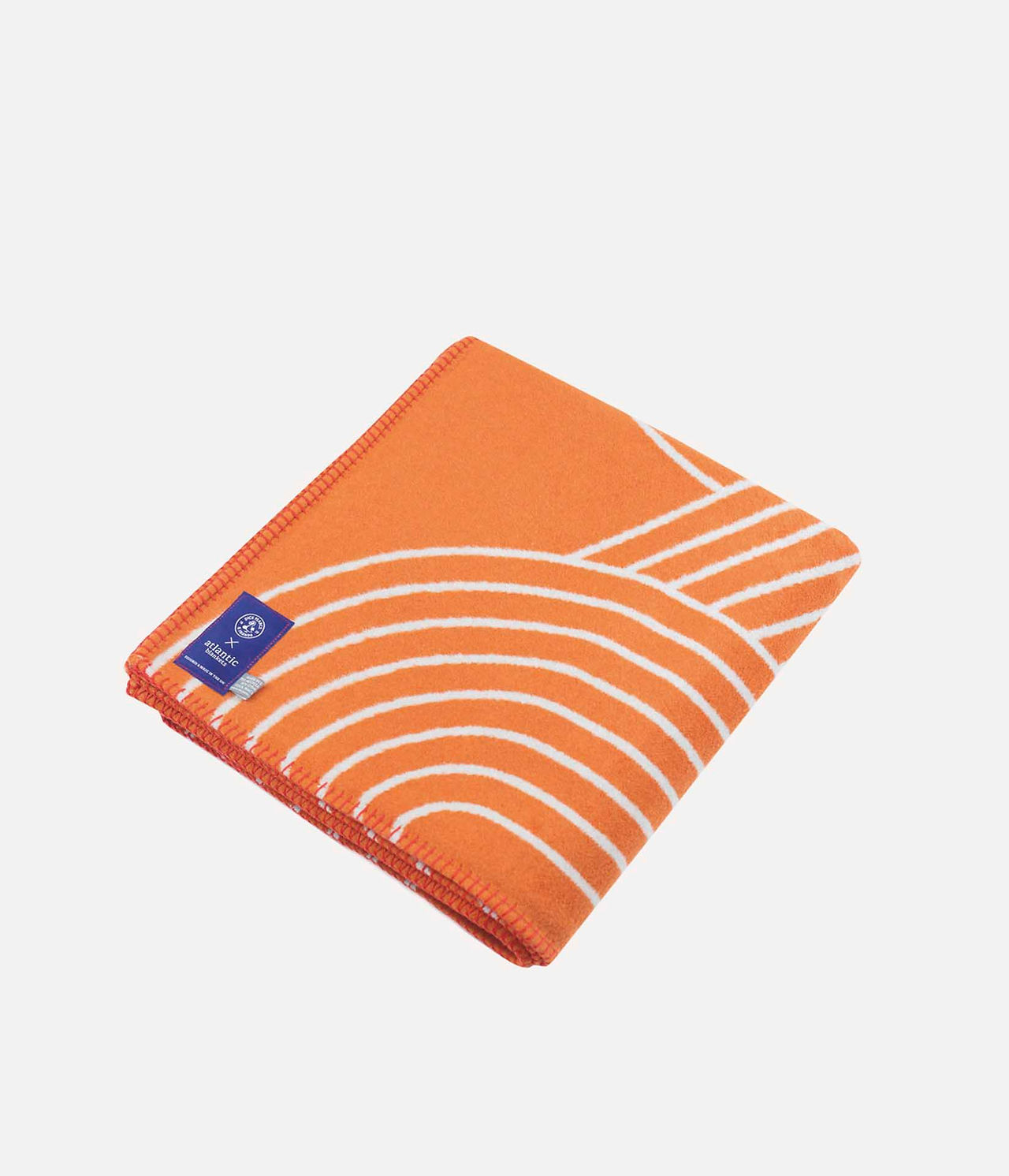 Folded orange blanket with white curved stripes on a white background, labeled Atlantic on a blue tag.