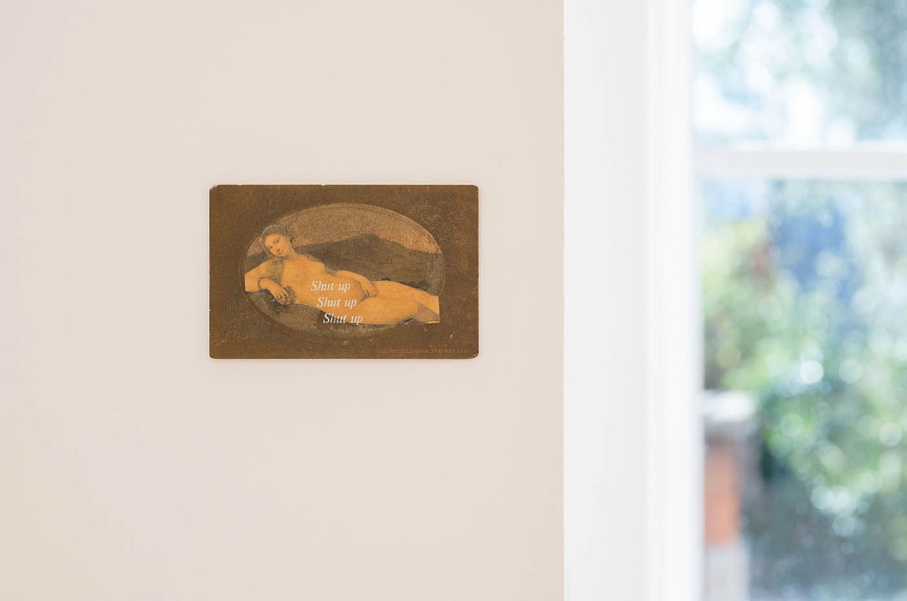 Small wall art of a reclining nude with Shut up repeated, hung on a beige wall beside a bright window.