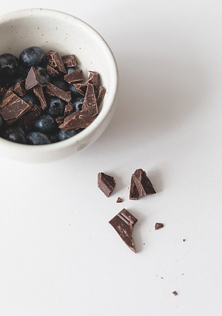 White bowl of blueberries and dark chocolate chunks on a white surface, with a few chocolate pieces scattered nearby.