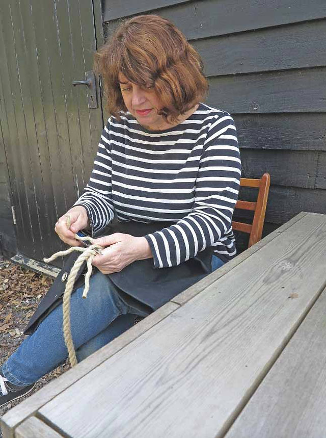 Woman in a black-and-white striped shirt sits outdoors, tying rope at a wooden table beside a dark wall.