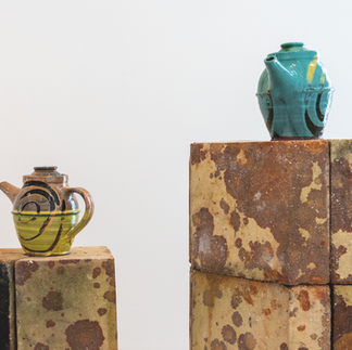 Two glossy ceramic teapots, one green and one turquoise, sit on rust-stained blocks against a plain white background.