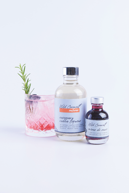 Pink cocktail with rosemary sprig beside two Wild Cornish liqueur bottles on a white background, labels reading autumn and crème de mure