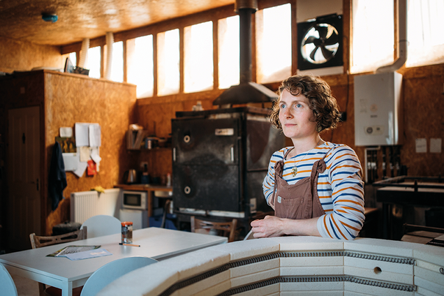   Woman in striped shirt and apron stands in a rustic workshop with a kiln and fan, looking thoughtfully off-camera.
