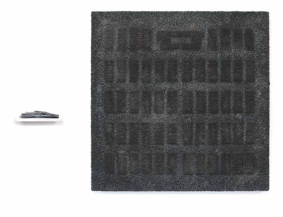Abstract art installation with a small folded gray piece on the left and a large dark textured square on a white wall.