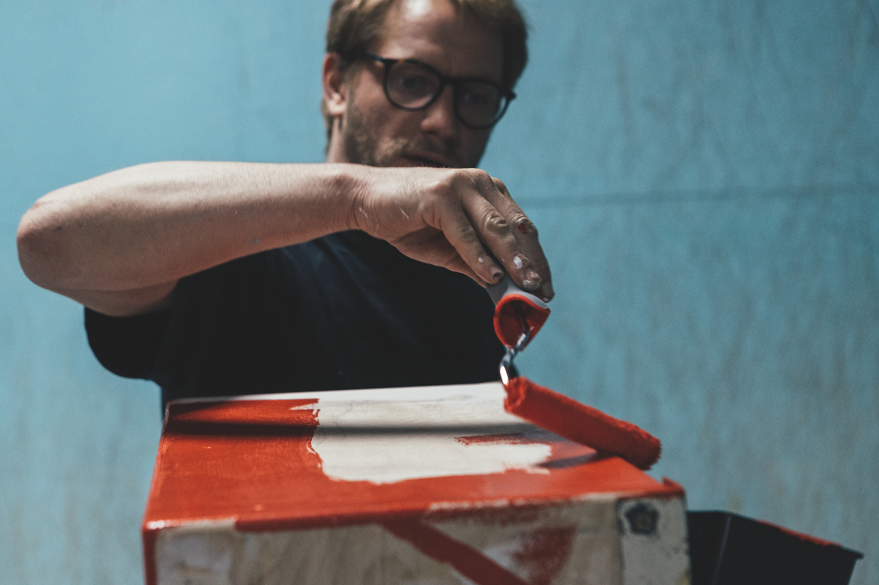 Man with glasses paints a box red and white with a roller in a blue room, focused and intent.