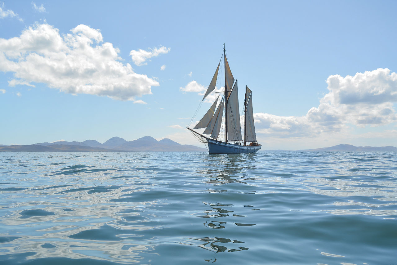 Sailboat with white sails glides on calm blue water under a sunny sky, with mountains and clouds in the background.