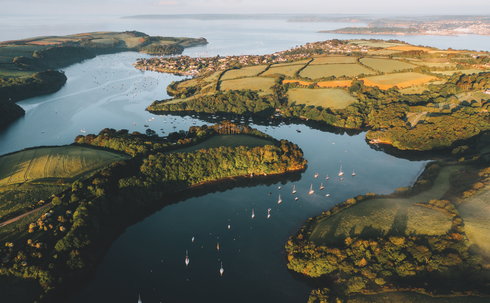 Aerial view of a calm bay with sailboats, green patchwork fields, wooded shores, and a coastal town in warm sunrise light. Image by photographer Joe Jackson.