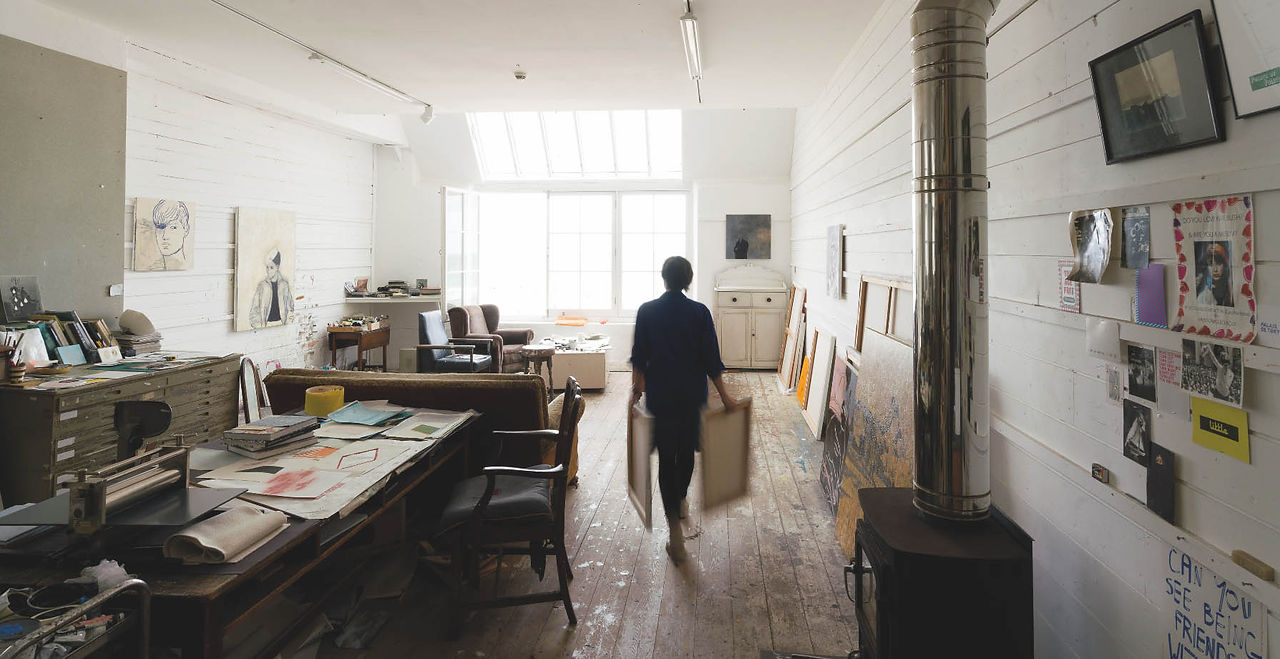 Person carrying framed canvases through a bright, cluttered artist studio with sketches, paintings, and a metal stove.