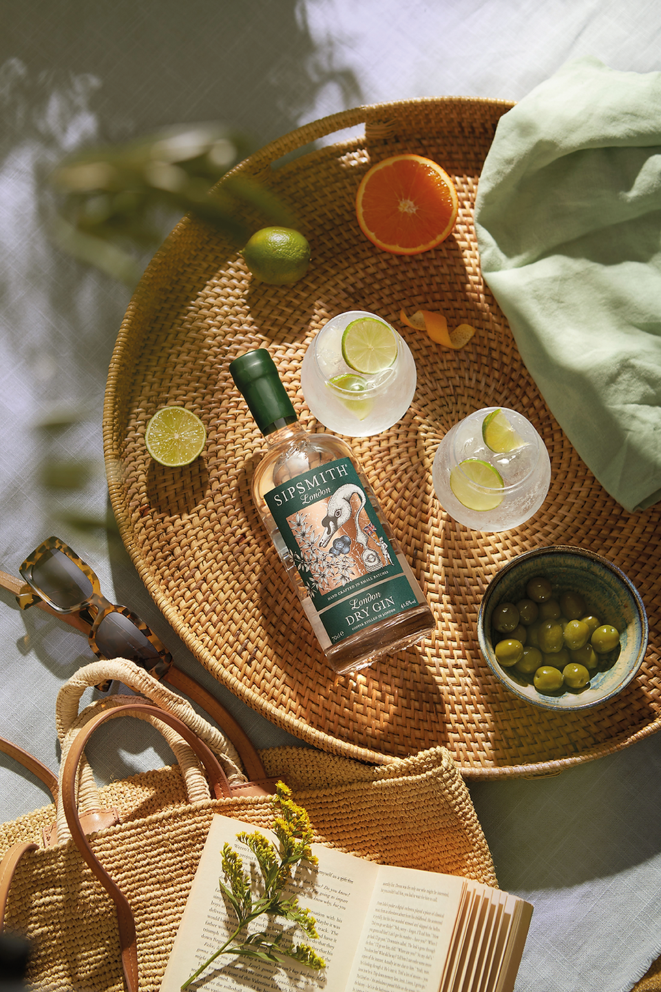 Sunlit picnic tray with Sipsmith London Dry Gin, citrus slices, olives, sunglasses, book and woven bag on a pale blanket.
