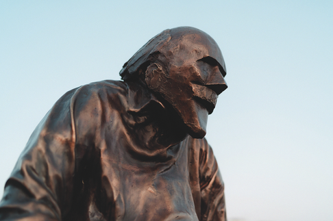 Bronze statue of a hooded man in profile against a pale sky, his worn face and shoulders lit warmly.