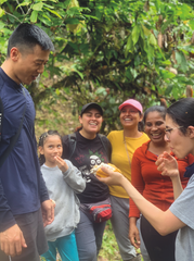 Smiling group outdoors in lush greenery, sharing a snack; a woman offers food while others laugh and watch.