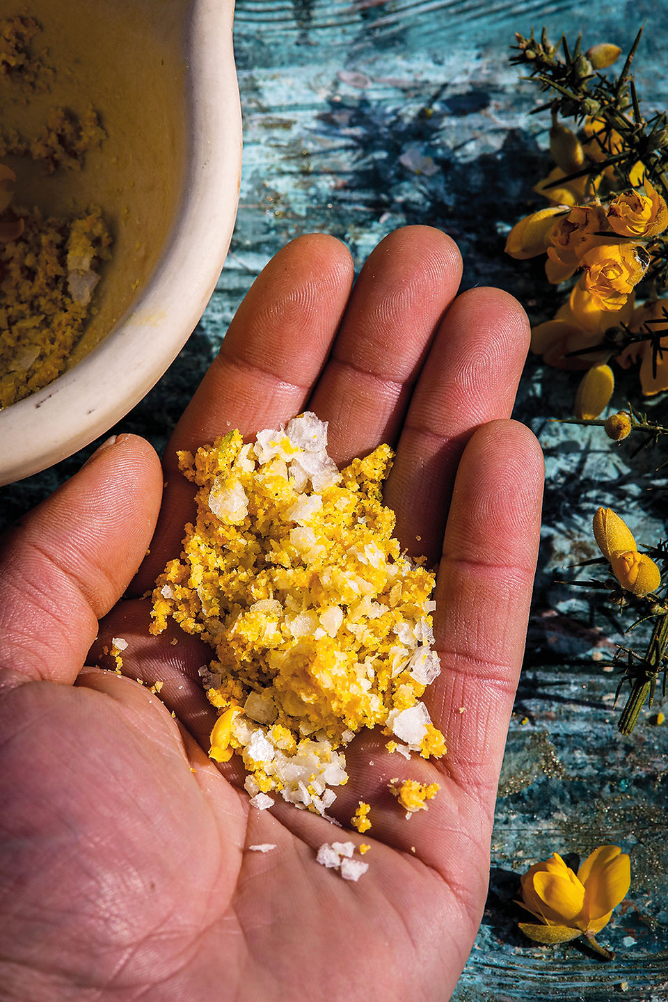 Hand holding yellow crumbly mixture beside a bowl on a blue rustic table, with yellow flowers in the background.