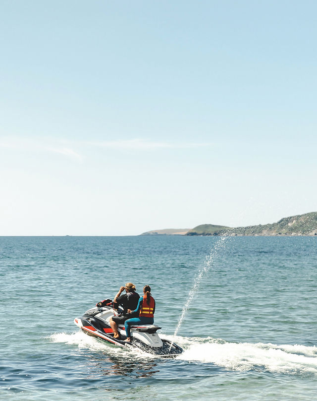   Two people ride a jet ski across calm blue sea, spraying water, with a distant coastline under a clear sky.