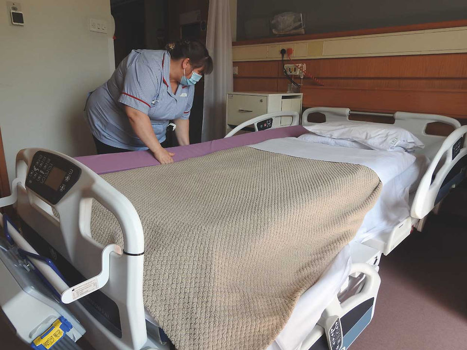 Nurse in a blue uniform and mask straightens a hospital bed with beige blanket in a quiet patient room.