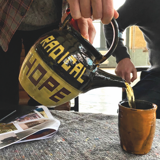Hands pour tea from a yellow-and-black RADICAL HOPE pot into a brown cup on a carpeted table, with a magazine nearby.