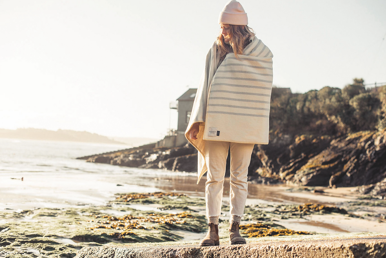 Woman in a pink beanie stands on a rocky beach, wrapped in a striped blanket, with a sunlit coast and house behind her. Blankets woven by Atlantic Blankets