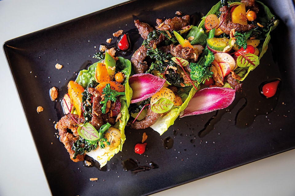 Three colorful beef lettuce wraps on a black plate, topped with vegetables and herbs, drizzled with sauce on a clean tabletop.