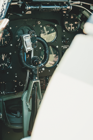 Vintage aircraft cockpit with black gauges and a control yoke; white seat edge in foreground.