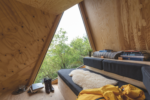 Wooden A-frame cabin nook with cushioned bench, books, boots and a yellow jacket by a triangular window to green trees.