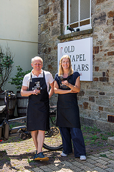 Two women in black aprons stand outside a stone building beside a sign reading OLD CHAPEL CELLARS, smiling with cameras.