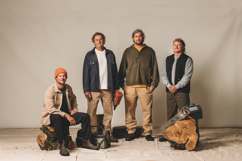  Four men pose in a studio on log stumps, one in an orange beanie seated at left, with a chainsaw and tool bag; calm, rugged.