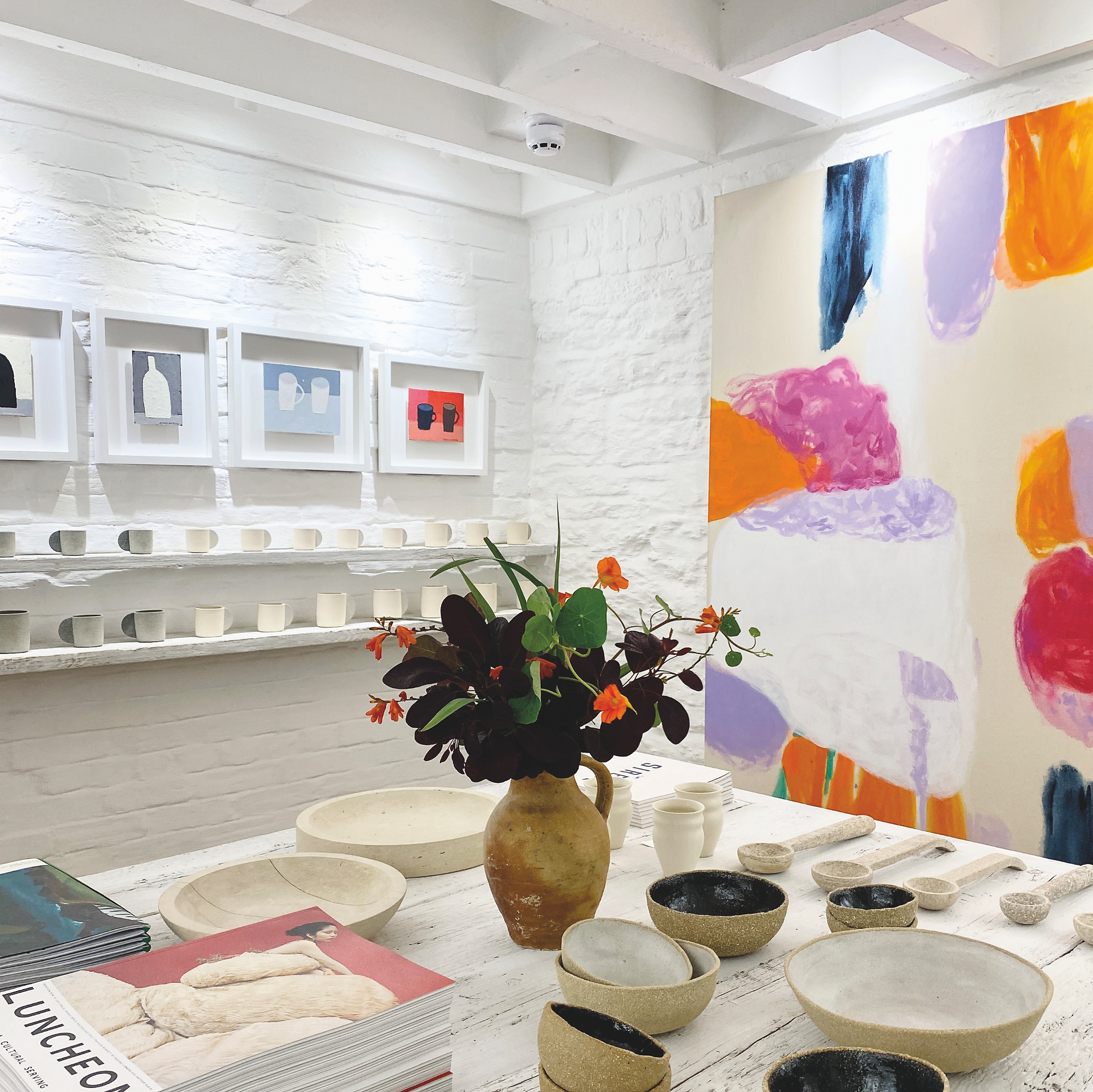 Bright art gallery with pottery on a white table, flowers in a vase, framed minimalist prints, and a colorful abstract mural on the wall at Morgans of Falmouth