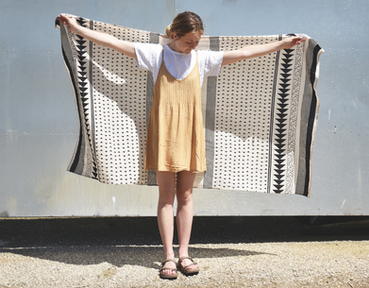 Young woman in a tan dress and white shirt holds out a black-and-white patterned cloth against a metal wall in bright sun