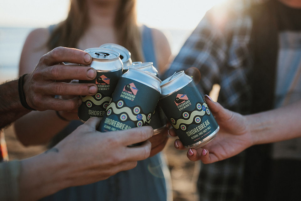 People clink Shorebreak Brewing cans at a beach sunset, with Firebrand Brewing text visible.