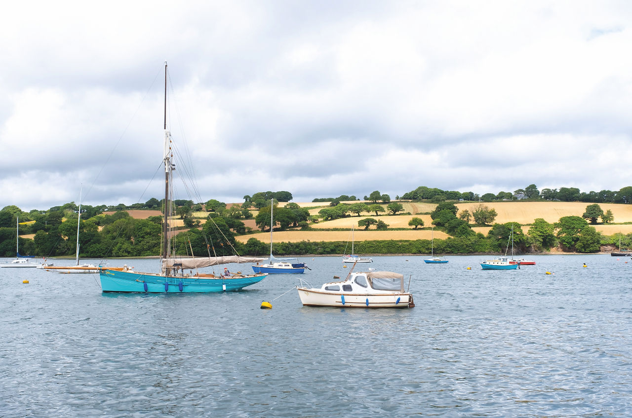 Sailboats and small moored boats on calm bay water, with green hillside fields and a cloudy sky.