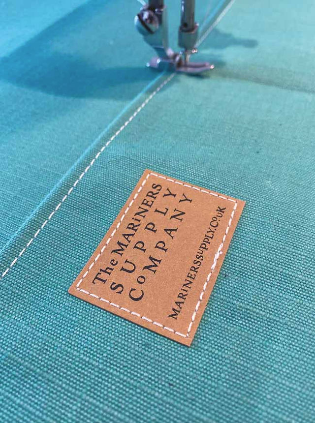 Close-up of a sewing machine stitching teal fabric with a leather label reading The Mariners Supply Company.