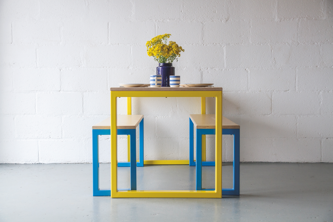 Yellow-framed table with blue stools, set with striped mugs, plates, and a vase of yellow flowers against a white wall. Designs by Coast Creative