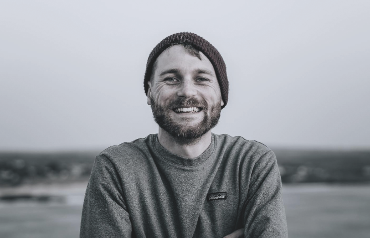 Smiling bearded man in a beanie and gray Patagonia shirt outdoors with a blurred landscape behind him. Photographer Joe Jackson.