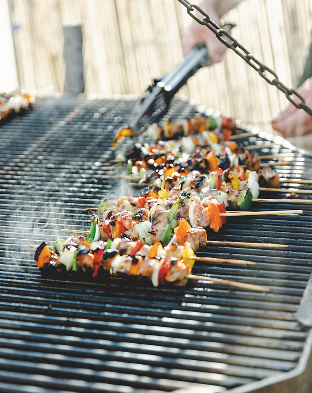  Skewers of grilled vegetables and meat sizzle on a barbecue as a person uses tongs, with smoke rising in an outdoor setting.