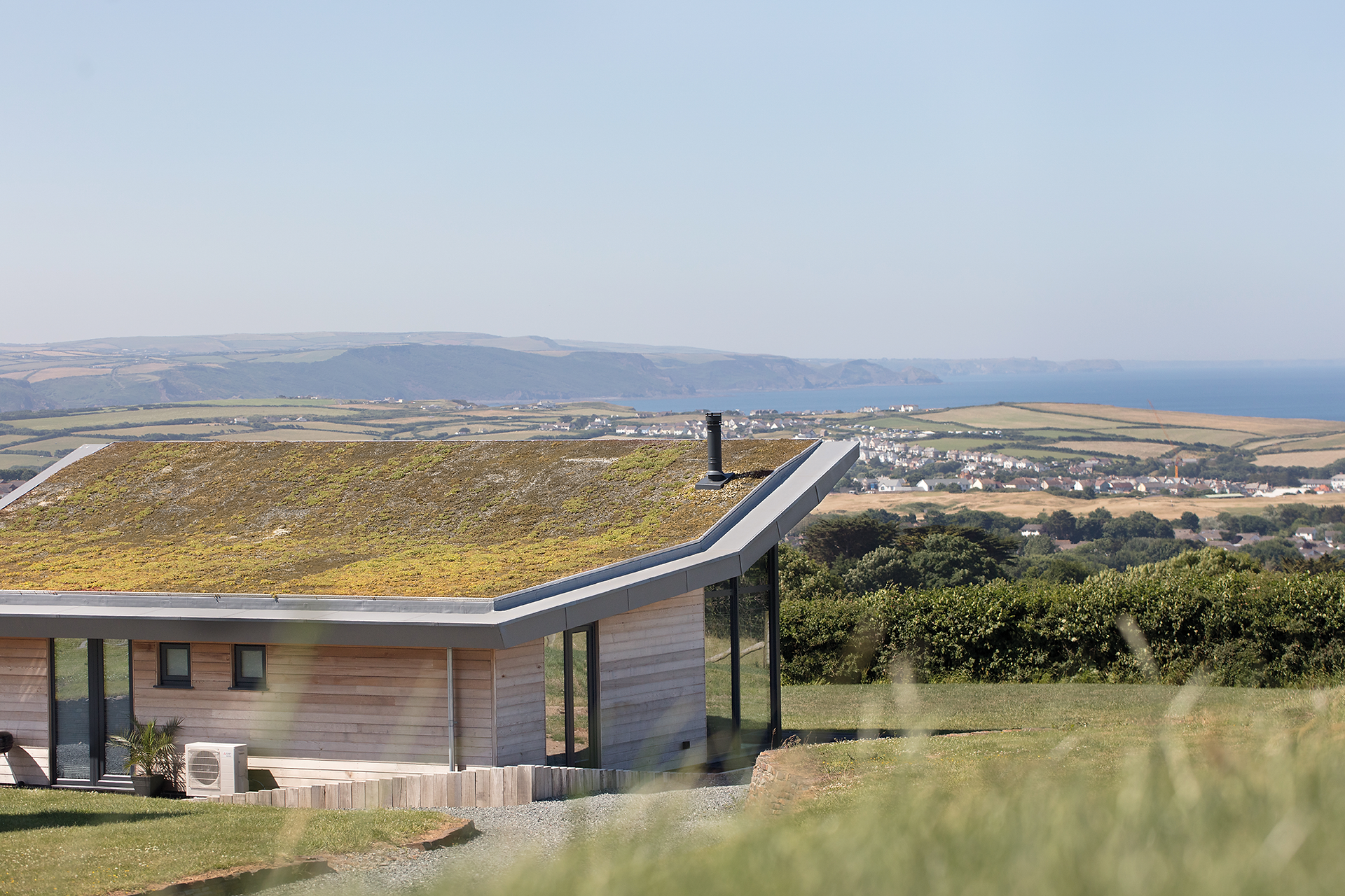 Modern house with a moss-covered roof overlooking coastal fields, village, and blue sea under a hazy sky