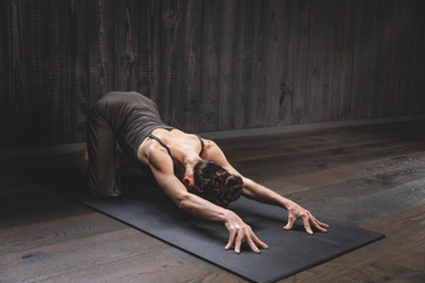 Woman in dark workout clothes in child’s pose on a black mat in a dim wooden yoga studio, calm and focused.