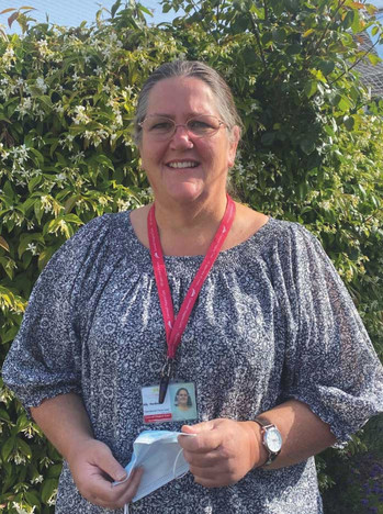 Smiling woman outdoors before a flowering bush, holding a face mask, wearing glasses, a lanyard, and an ID badge.