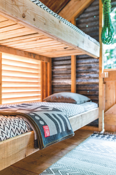 Rustic wooden bunk bed with patterned blanket and pillow in a sunlit cabin, with a red label on the blanket.