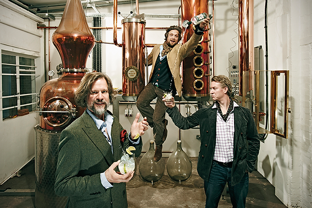 Three men pose in a distillery with copper stills; one stands on glass holding a drink, another raises a bottle, and one holds a cocktail.