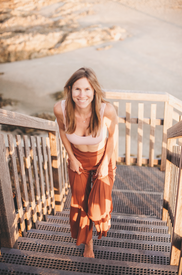Smiling woman in a pink top and rust skirt climbs outdoor stairs by a rocky beach in warm sunlight.