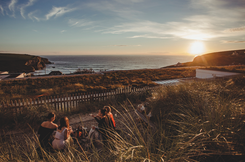 Group of women sharing drinks on a grassy seaside slope at sunset, with cliffs, ocean, and glowing sky. from the rooftop terrace at the Bedruthan Hotel and Spa