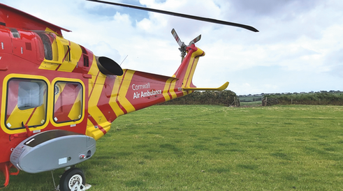 Red and yellow Cornwall Air Ambulance helicopter parked on a grassy field under a cloudy sky.
