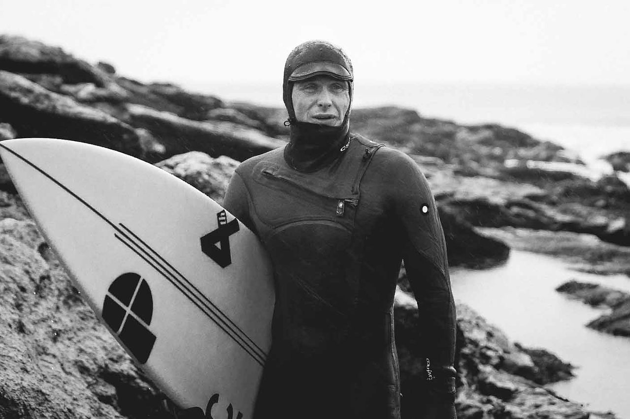 Black-and-white surfer in a wetsuit holds a surfboard on a rocky shoreline, facing the camera with a focused expression. By Photographer Luke Gartside 