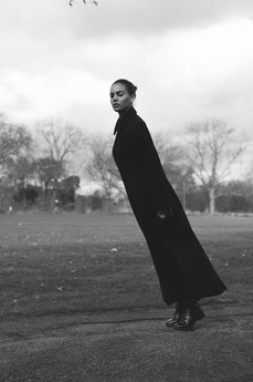 Black-and-white portrait of a woman in a long black coat and boots leaning in a park under a cloudy sky, looking serious.
