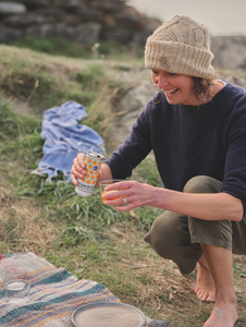 Smiling woman in a knit hat pours a drink from a patterned can into a glass during an outdoor picnic on grass.